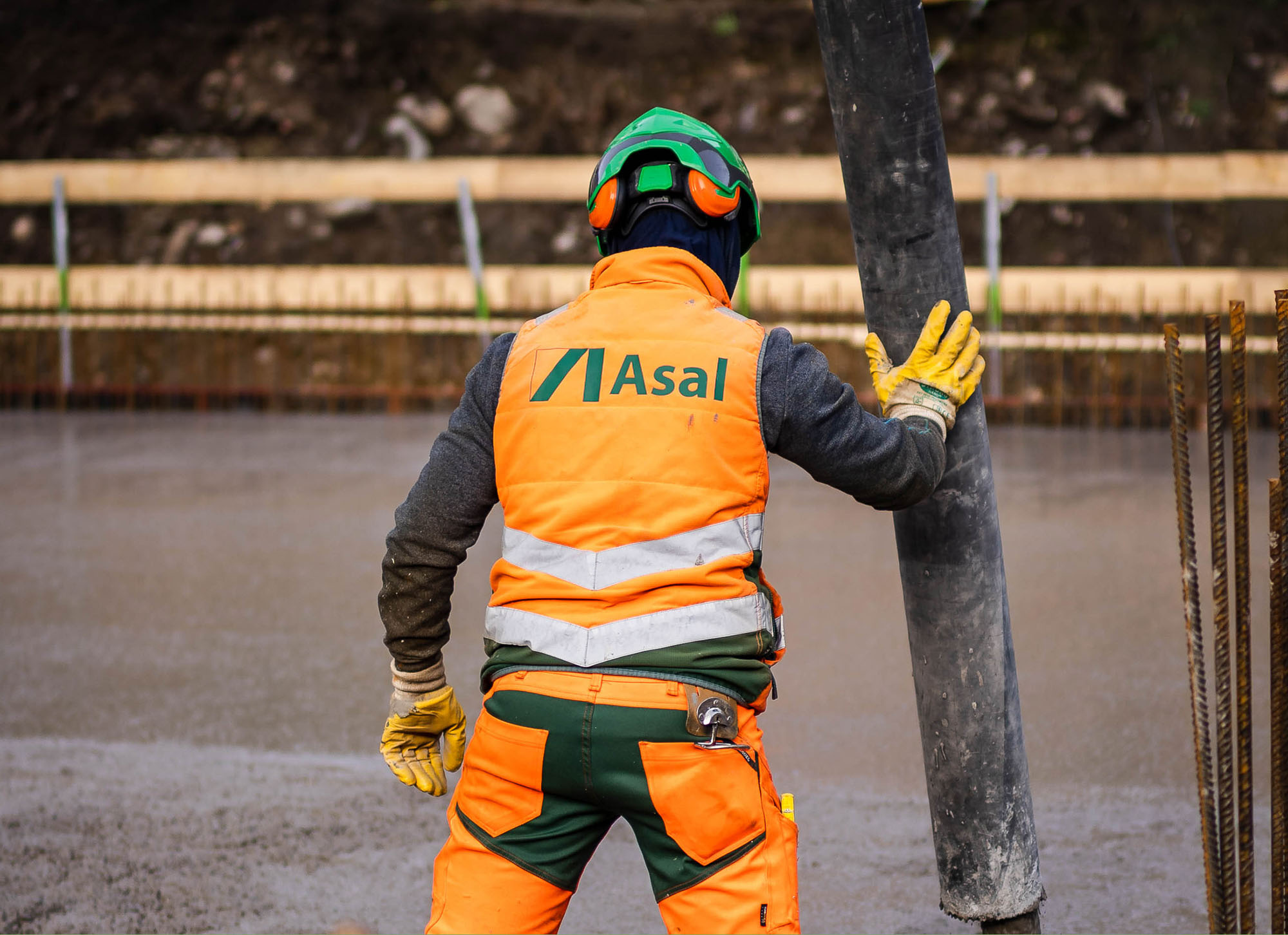 Zwei Bauarbeiter in orangefarbener Schutzkleidung und grünen Helmen auf einer Baustelle, im Hintergrund Holz und Betonwände.