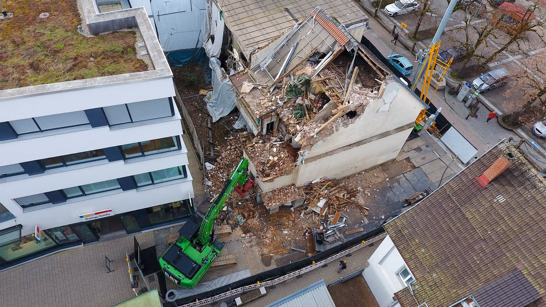 Aus der Vogelperspektive ist ein teilweise abgerissenes Gebäude zu sehen, dessen Dach eingestürzt ist und dessen Innenräume offenliegen. Ein grüner Bagger arbeitet zwischen Trümmern, während das Gebäude von einer Straße und benachbarten Häusern umgeben ist.