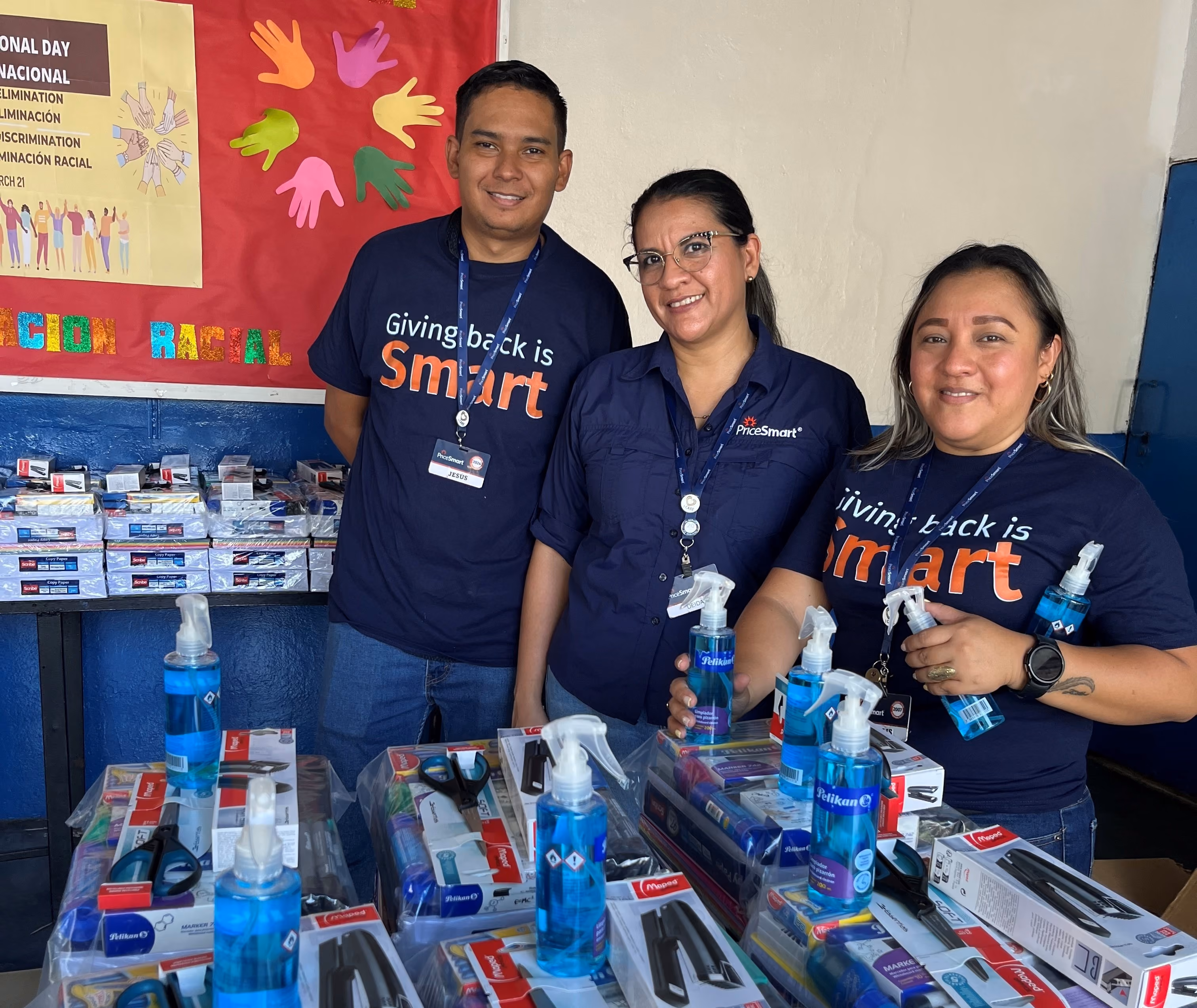 Three volunteers wearing PriceSmart shirts stand behind tables filled with school supplies and blue sanitizer bottles.