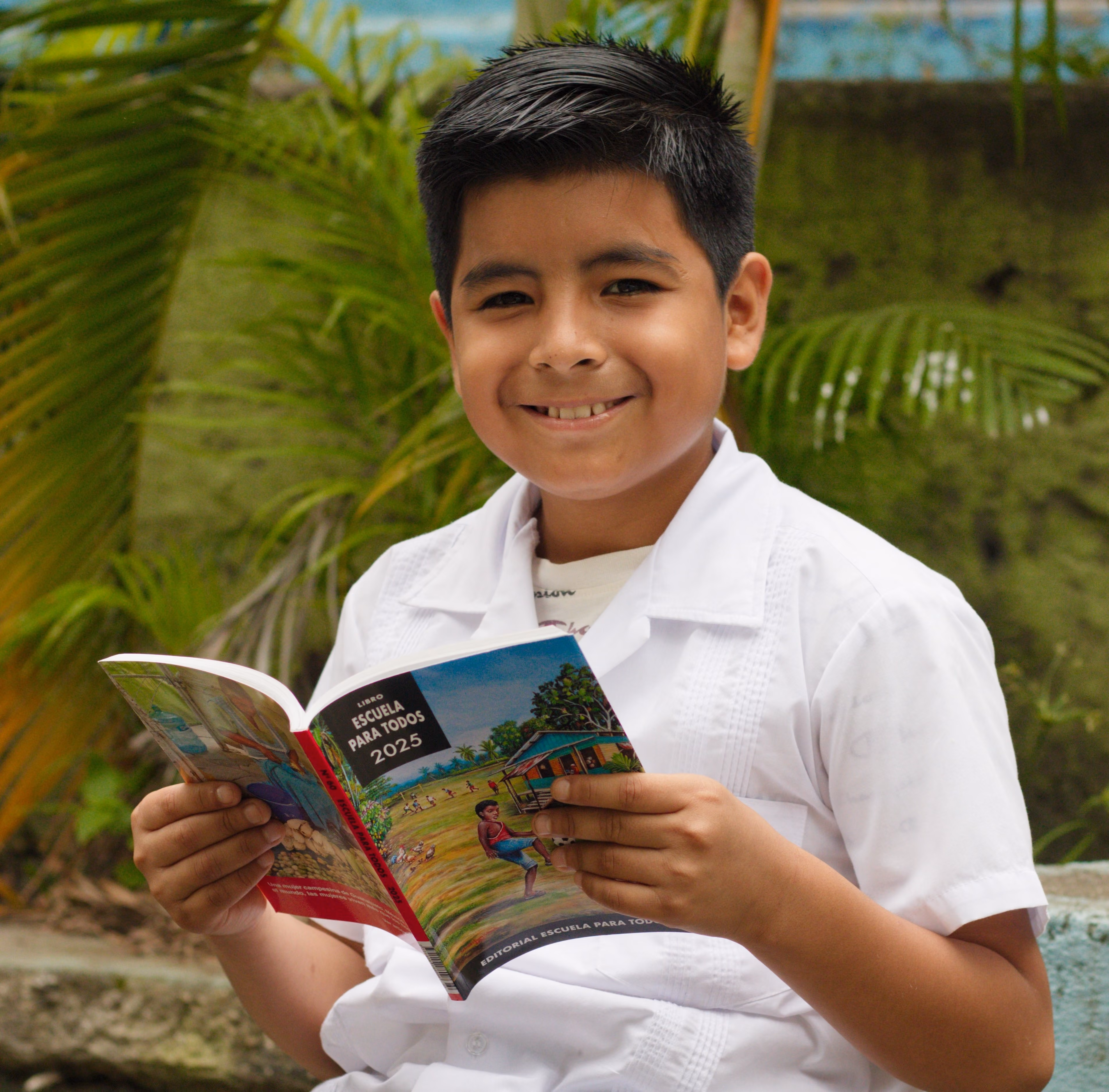 Smiling boy in white shirt holding and reading a colorful book outdoors with green plants background.