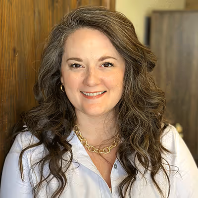 Smiling woman with long wavy hair wearing a white blouse and gold chain necklace in a warmly lit room.