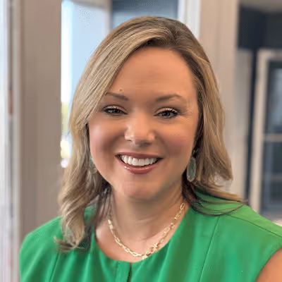 Smiling woman with blonde hair wearing a green top and gold necklace in a softly lit indoor setting.