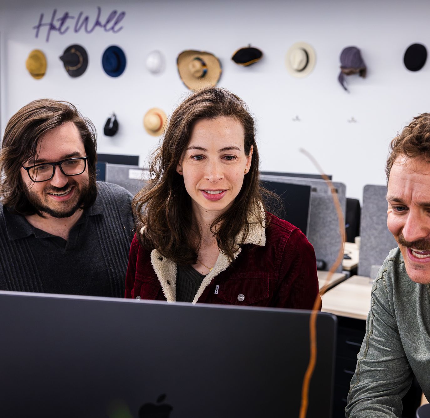 Three BigHat colleagues laughing together at a computer in front of the hat wall