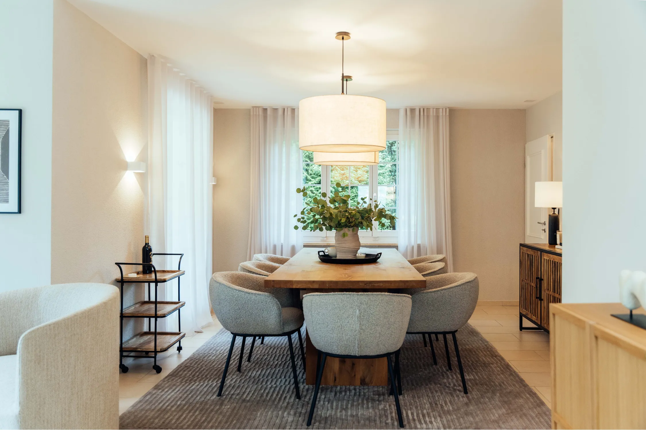 Modern dining room with a wooden table, grey chairs, hanging lamp, and white curtains.