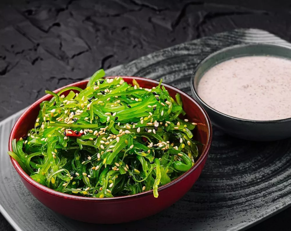 En rød skål fyldt med tangsalat toppet med sesamfrø står på en mørk tallerken ved siden af en lille skål med cremet dyppelse på en sort baggrund med struktur.