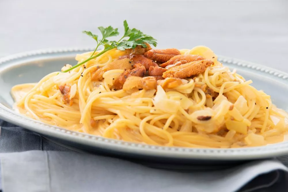 En tallerken med cremet pasta toppet med stykker af søpindsvin (uni) og pyntet med frisk persille, serveret på et lyseblåt fad med en grå serviet under.