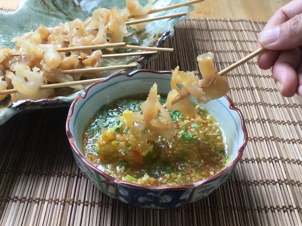 En hånd holder et spyd med vandmandsstykker dyppet i en skål med krydret sauce, med flere vandmandsspyd på en keramisk tallerken i nærheden, alt sammen på en bambusmåtte.
