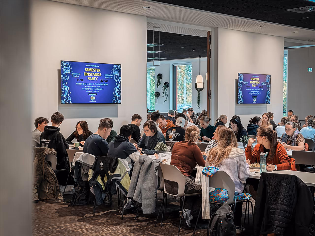Bildschirmwerbung auf dem Campus Ulmenstraße in Rostock. Studierende essen und schauen auf die Werbebildschirme. 