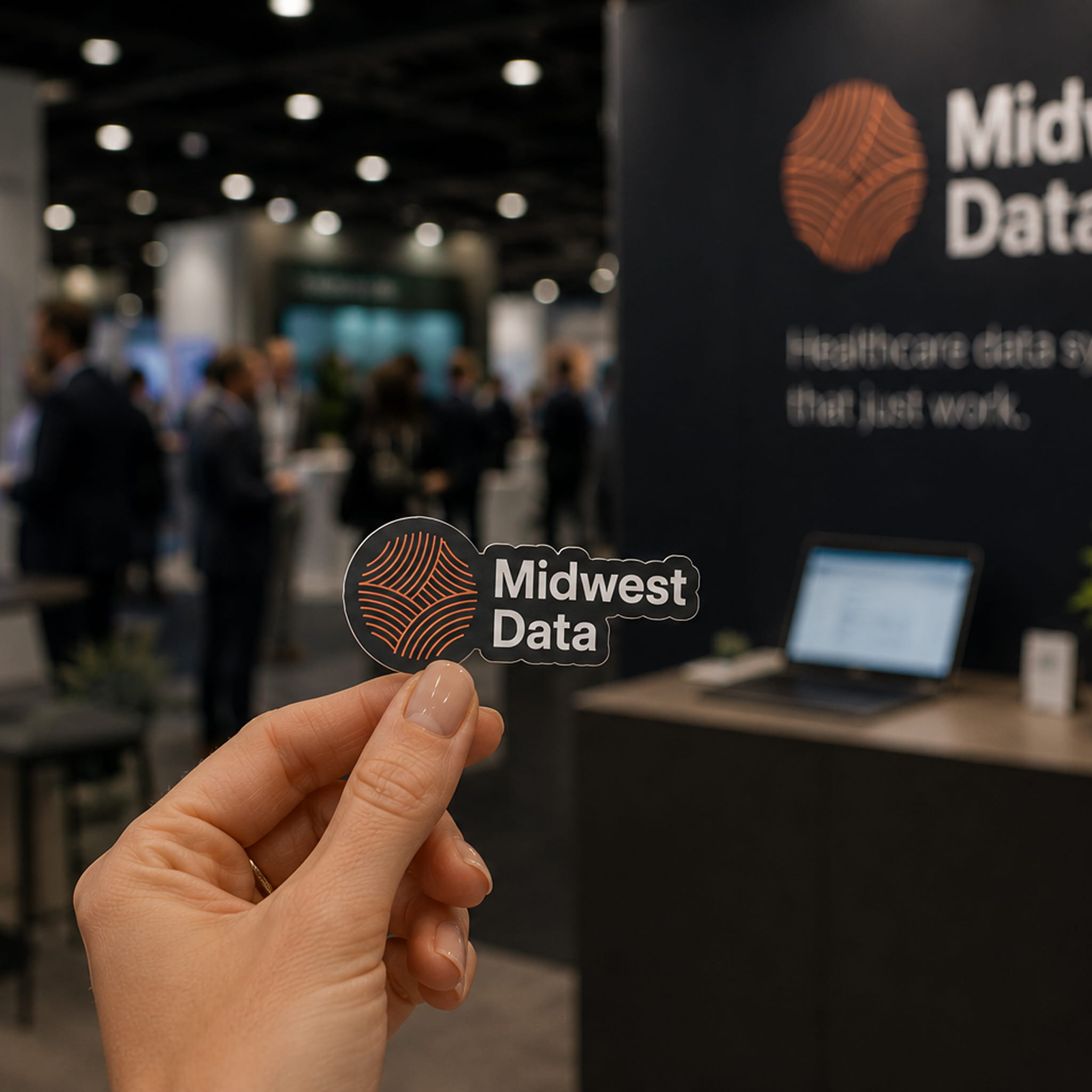 Hand holding a Midwest Data sticker with logo at a blurred event booth background.