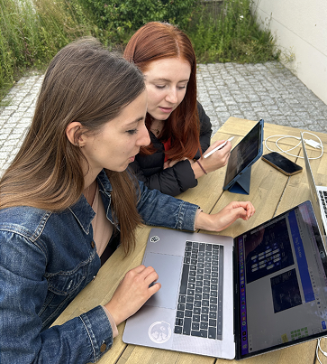 Two women sitting at a wooden outdoor table working on a laptop and a tablet, with a smartphone and laptop charging cables nearby.