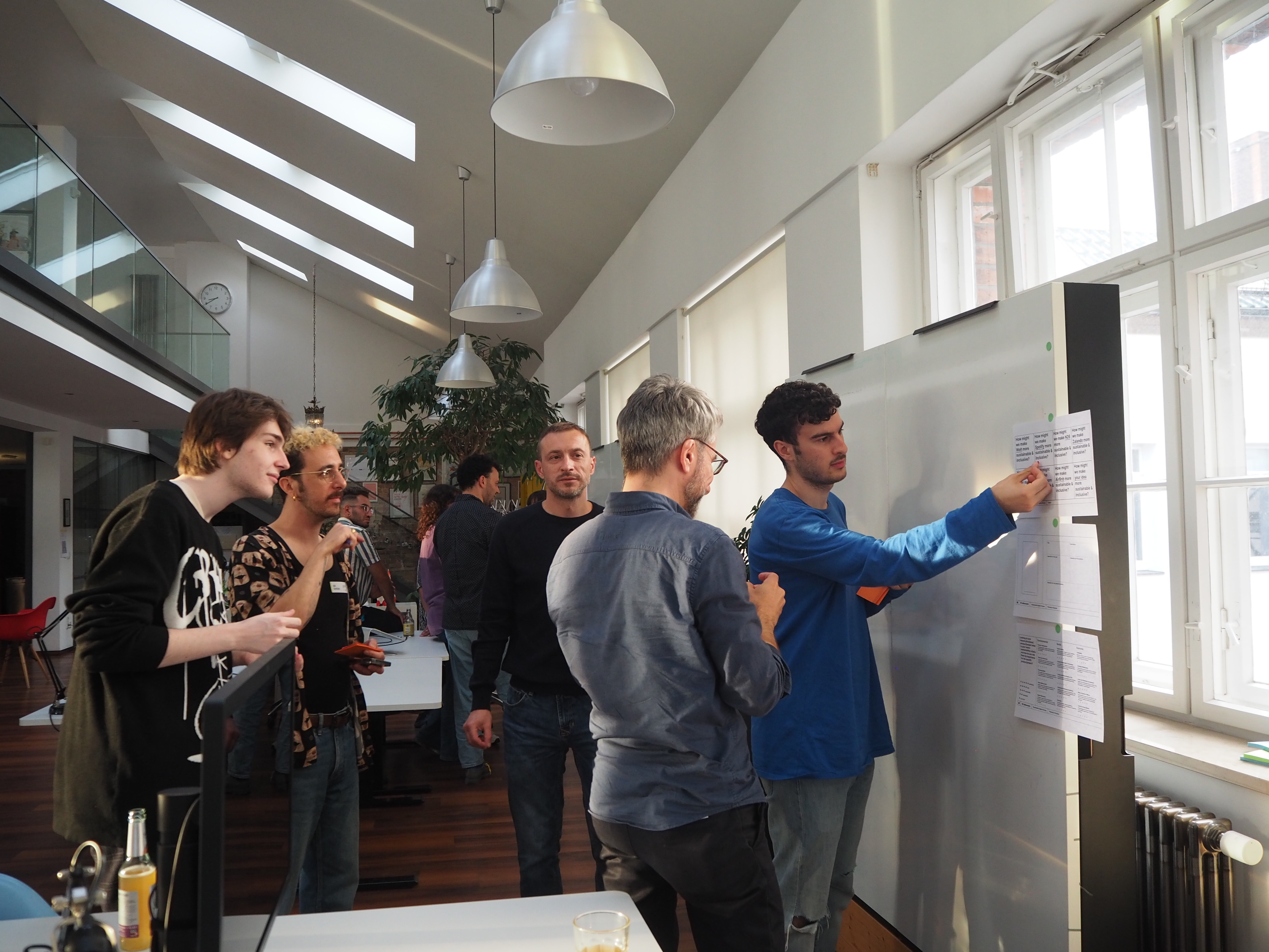 Group of people discussing and placing note papers on a whiteboard in a bright office with large windows and overhead lights.