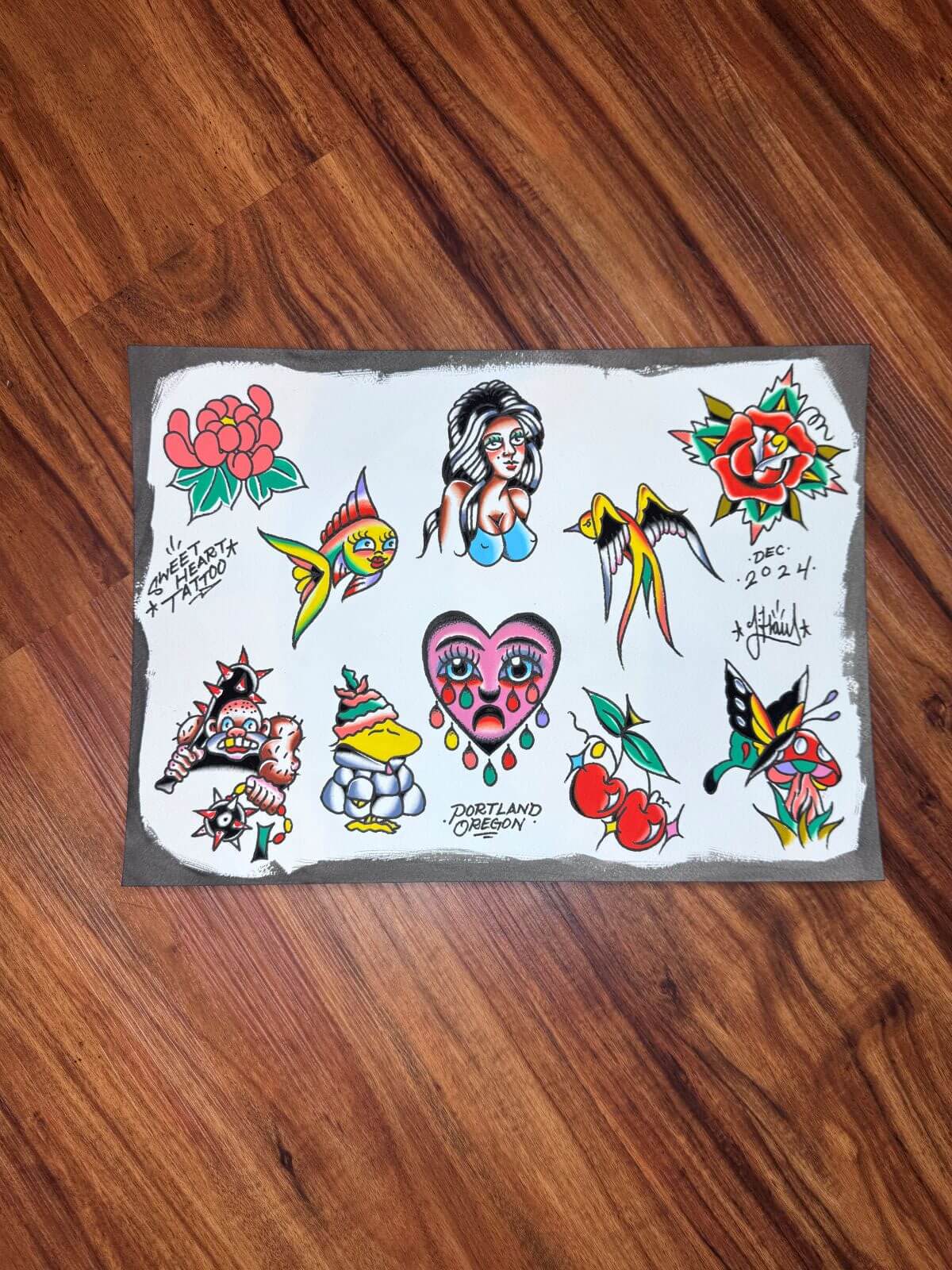 Tattoo flash sheet on wood floor featuring colorful traditional-style designs including a crying heart, a woman bust, birds, flowers, cherries, a mushroom with a butterfly, a fish, and a warrior helmet with spiked mace.