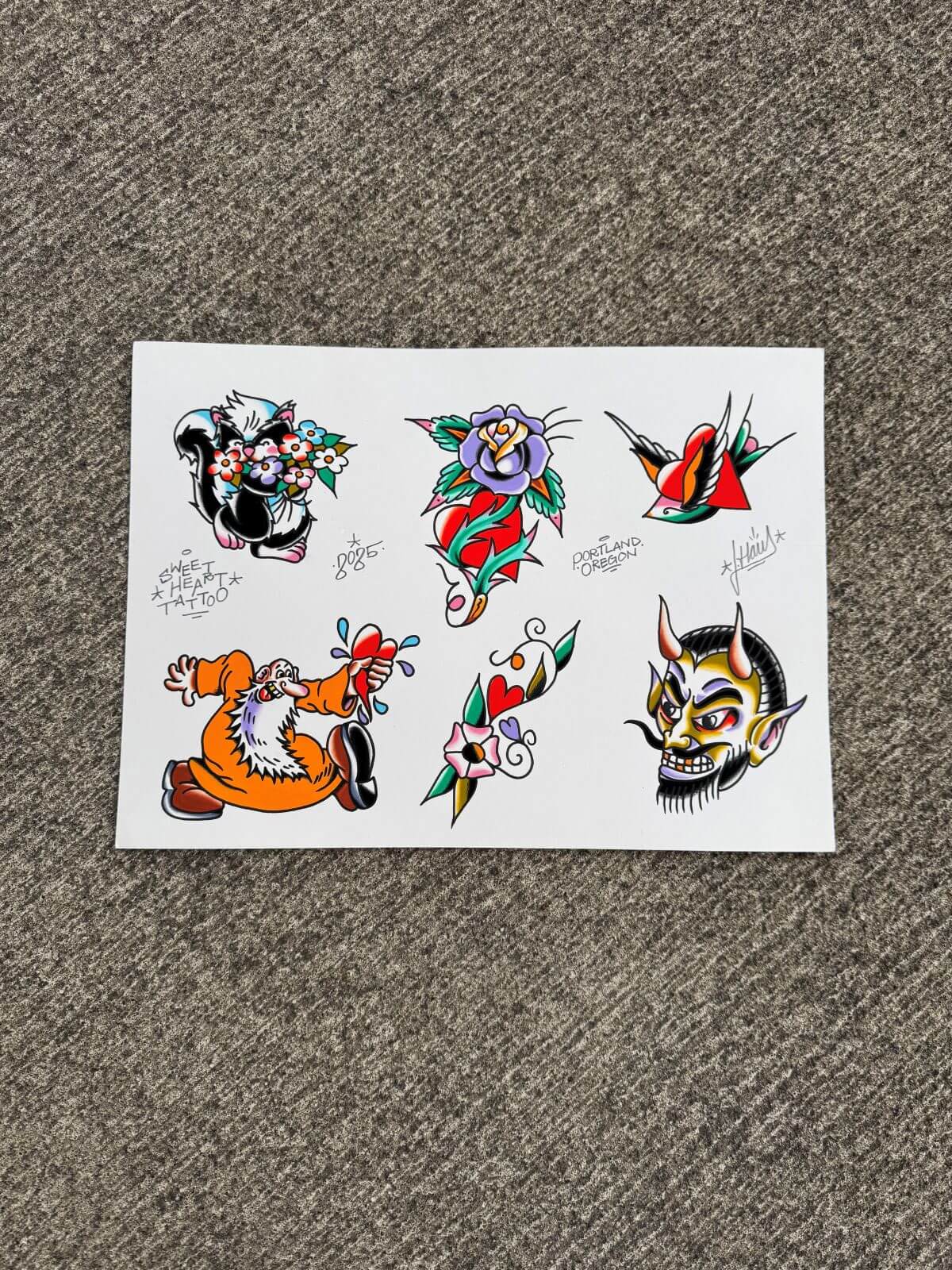 Traditional tattoo flash designs including a skunk with flowers, a purple rose with leaves, a swallow with a red heart, a dancing old man holding a bleeding heart, a dagger through a flower and heart, and a devil face with horns and a goatee.