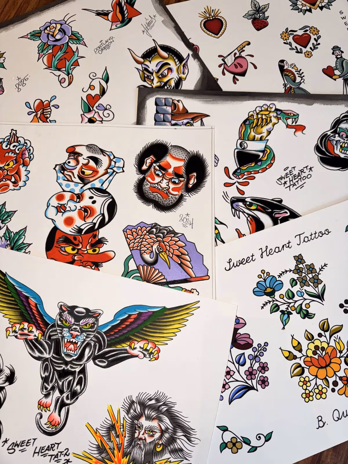 Collection of traditional flash tattoo designs including a winged black panther, stylized faces, a snake wrapped around a hand, flowers, hearts, and a crane on a fan.