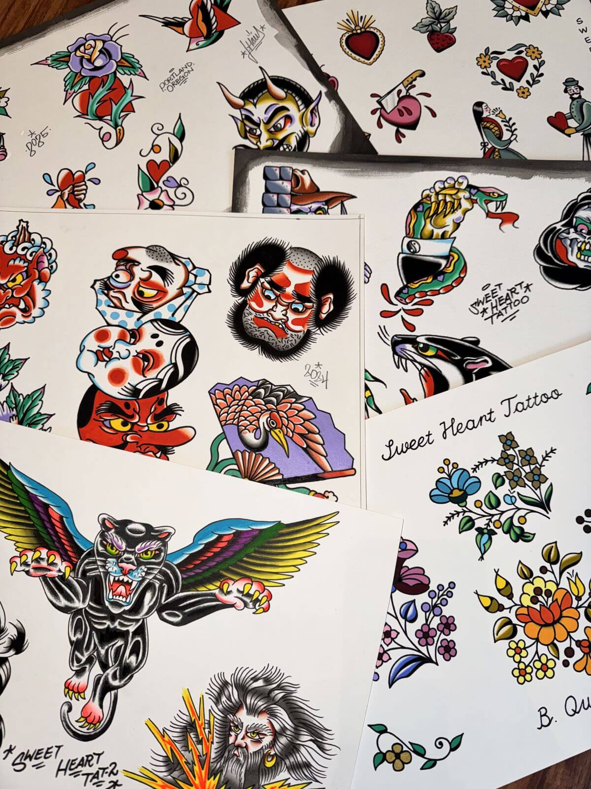 Collection of traditional flash tattoo designs including a winged black panther, stylized faces, a snake wrapped around a hand, flowers, hearts, and a crane on a fan.