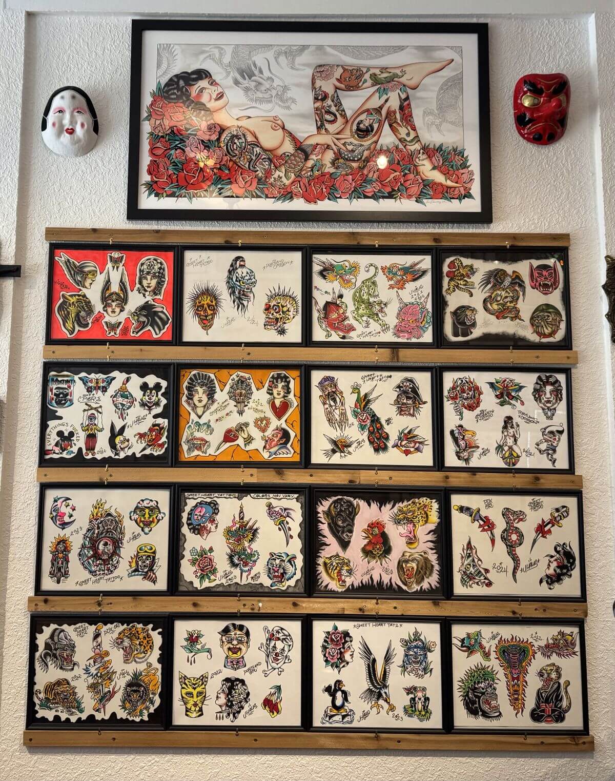 Wall display featuring a framed tattoo-style artwork of a tattooed woman lying on roses, flanked by two masks, above multiple framed tattoo flash sheets with traditional tattoo designs all done at Sweet Heart Tattoo.