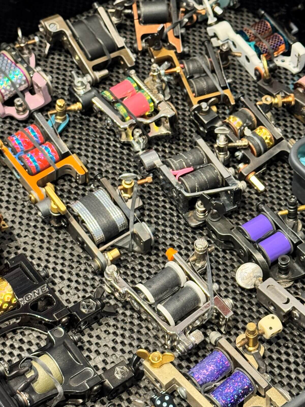 Arrangement of various colorful coiled tattoo machines placed on a textured black surface.