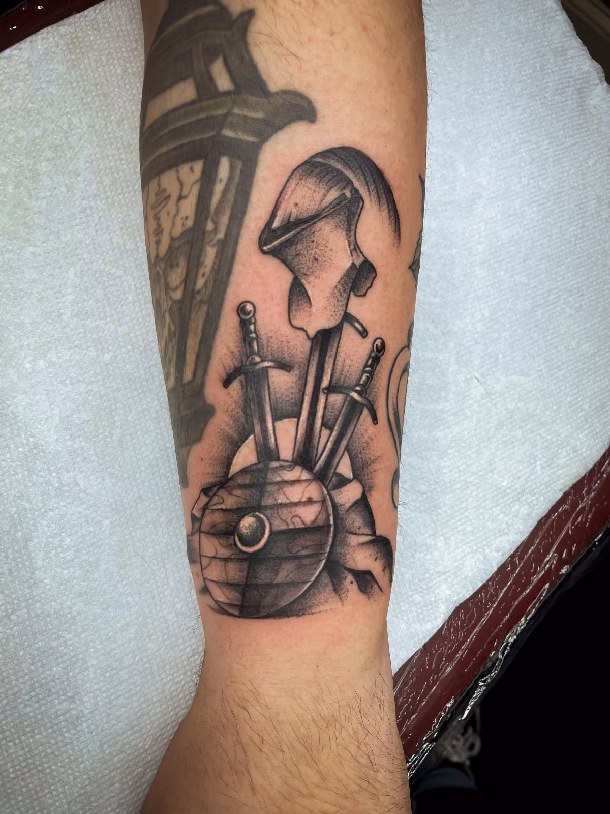 Black and grey tattoo of a medieval helmet, two swords, and a wooden shield on a forearm.