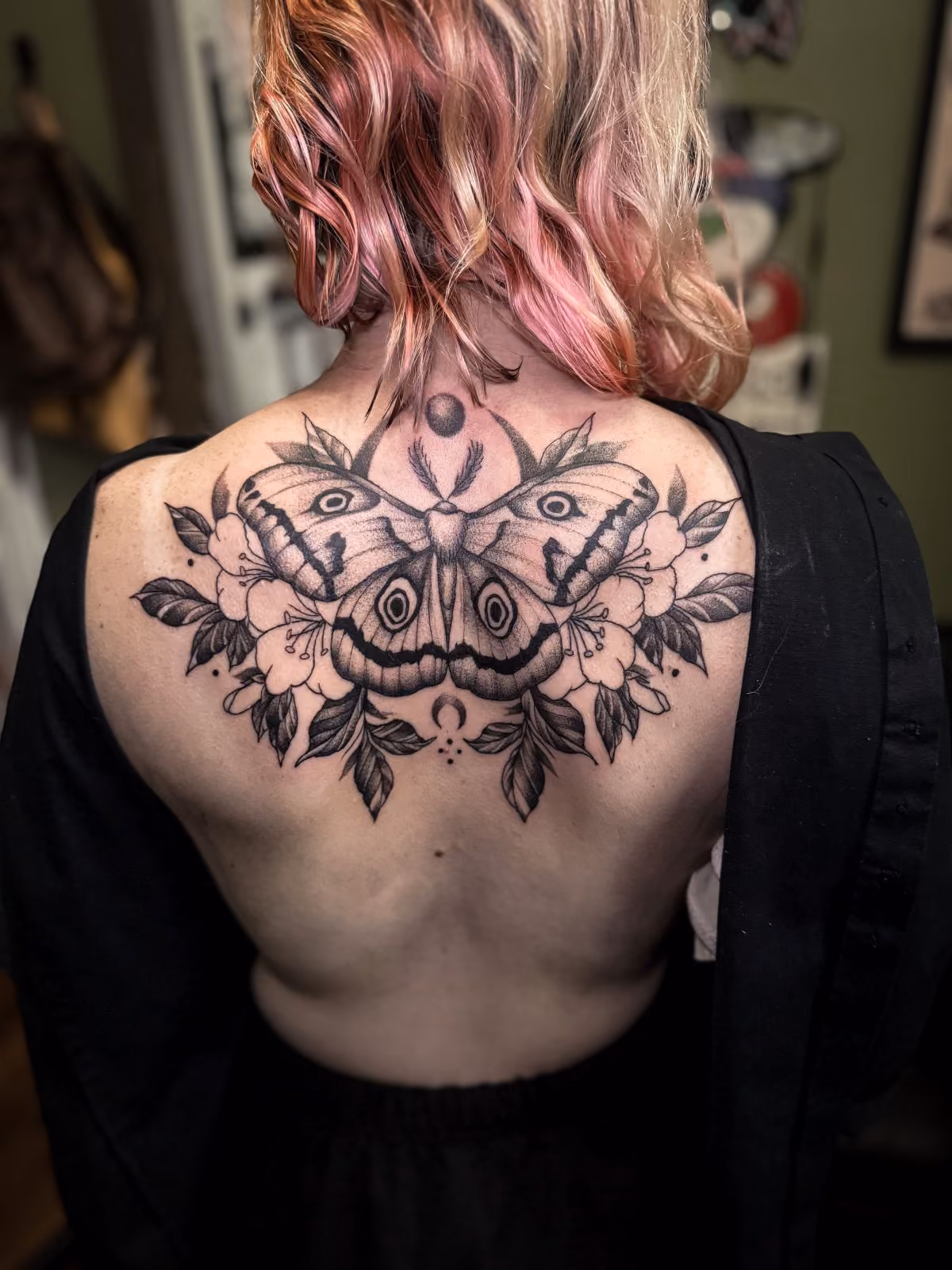 Black and grey tattoo of a large moth with detailed wings and surrounding flowers on upper back.