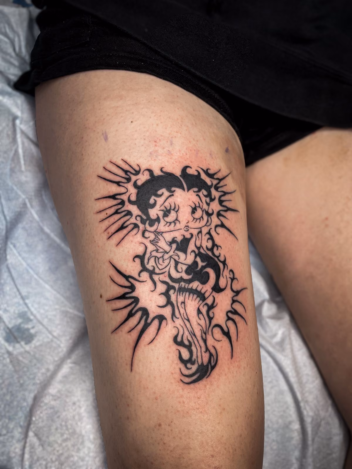 Black ink tribal-style tattoo of Betty Boop on a person's thigh.