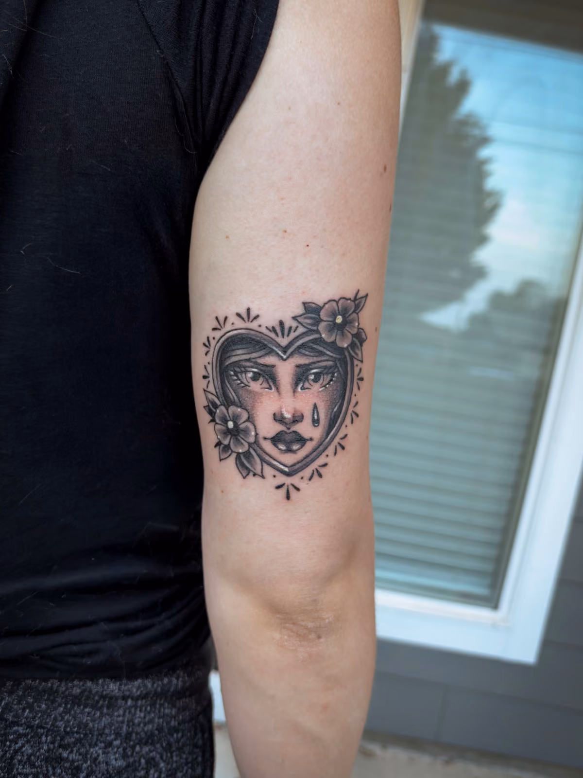 Black and grey tattoo of a woman's face with a tear inside a heart, decorated with flowers on arm.