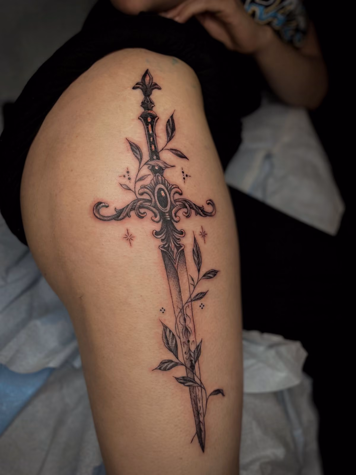 Tattoo of an ornate sword with leaves wrapping around the blade on a person's thigh.