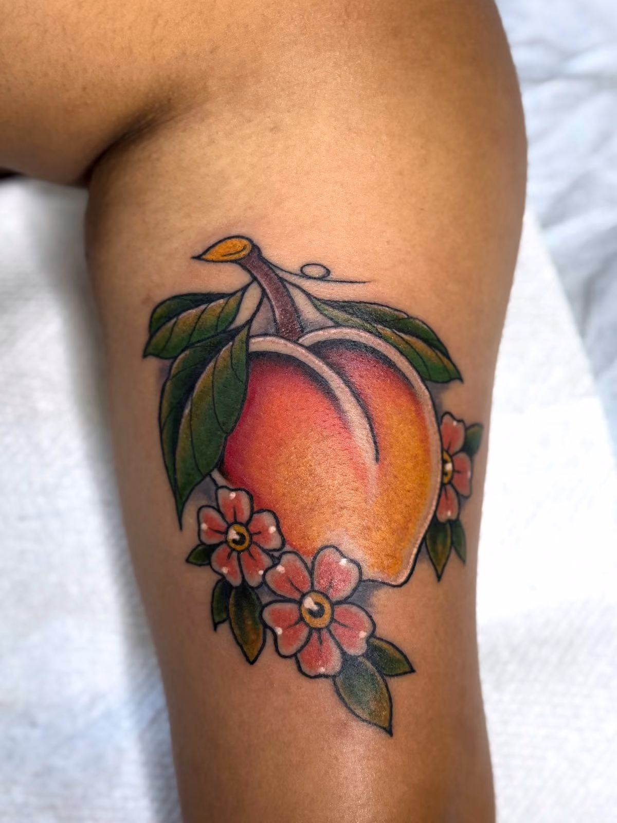 Tattoo of a peach with green leaves and pink flowers on a person's arm.
