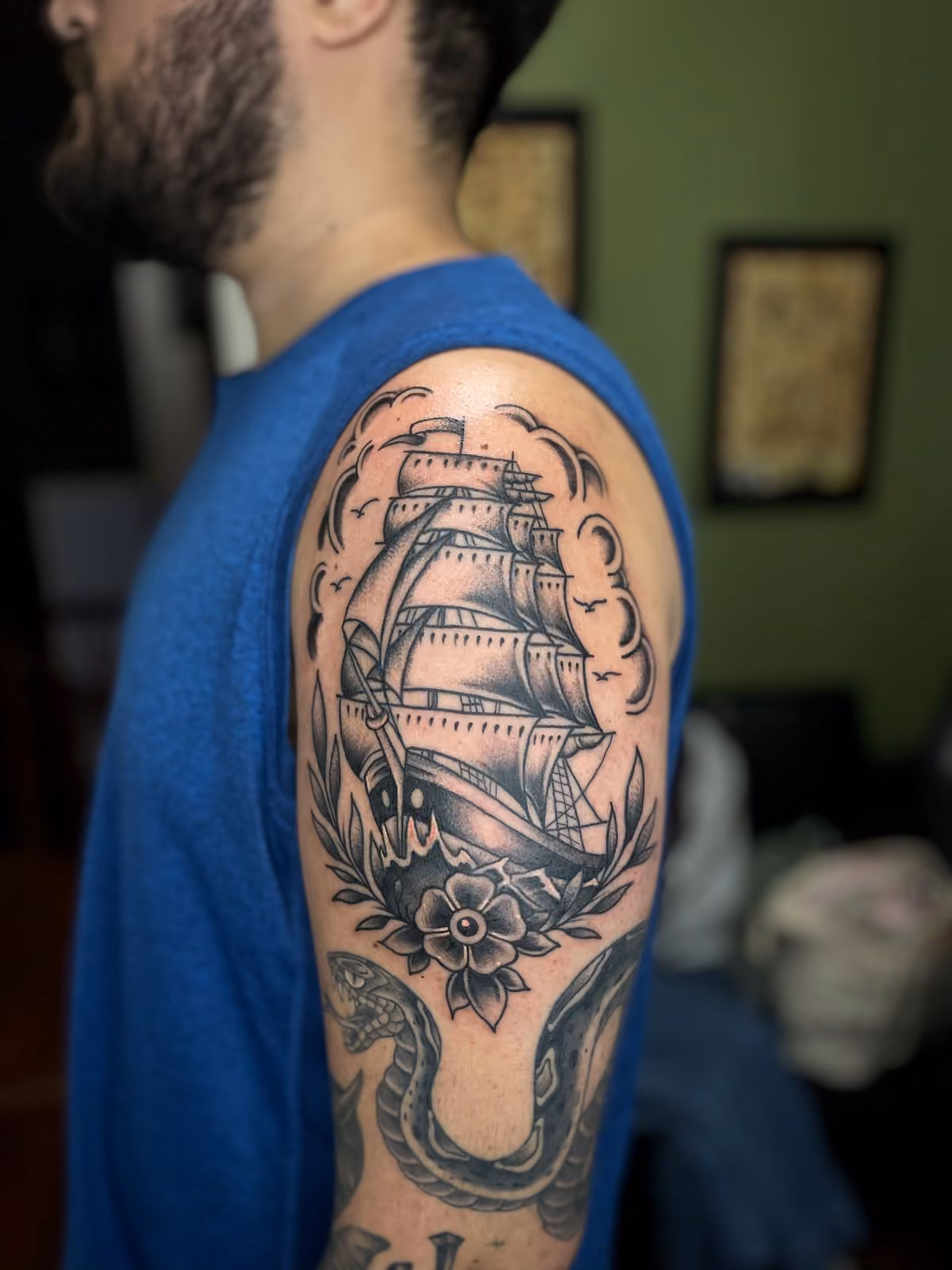 Black and gray tattoo of a traditional clipper ship, flower, and bird silhouettes on upper arm.