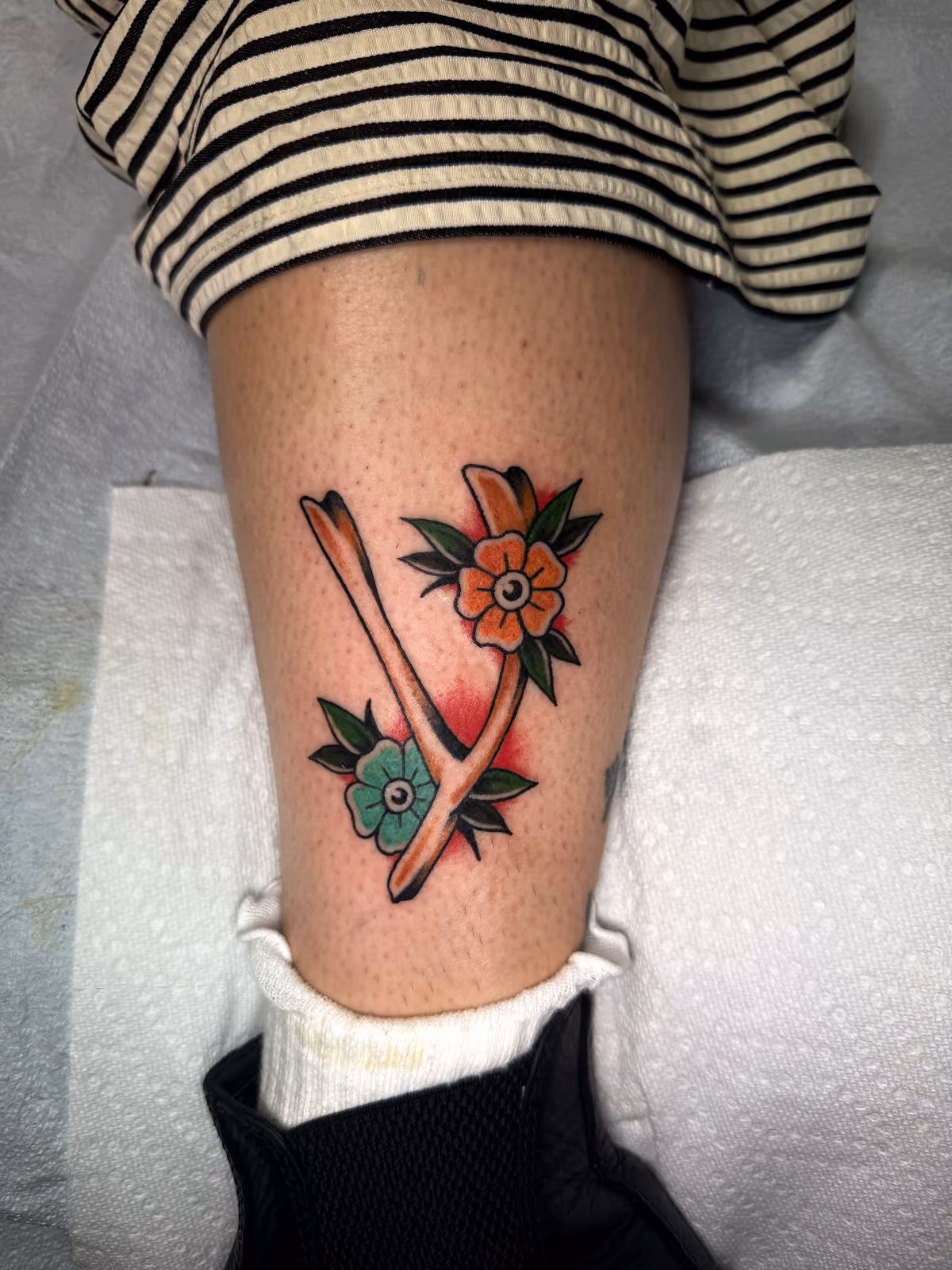 Traditional tattoo of a wishbone with orange and teal flowers and green leaves on a lower leg.