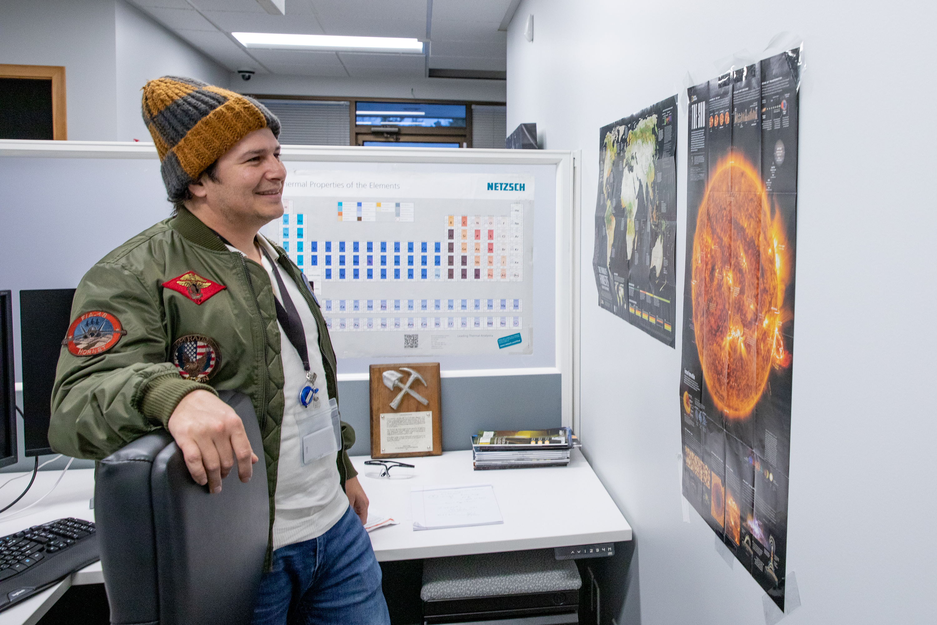 A man in a beanie cap smiles as he looks upon a poster of the sun at his desk at Zap Energy.