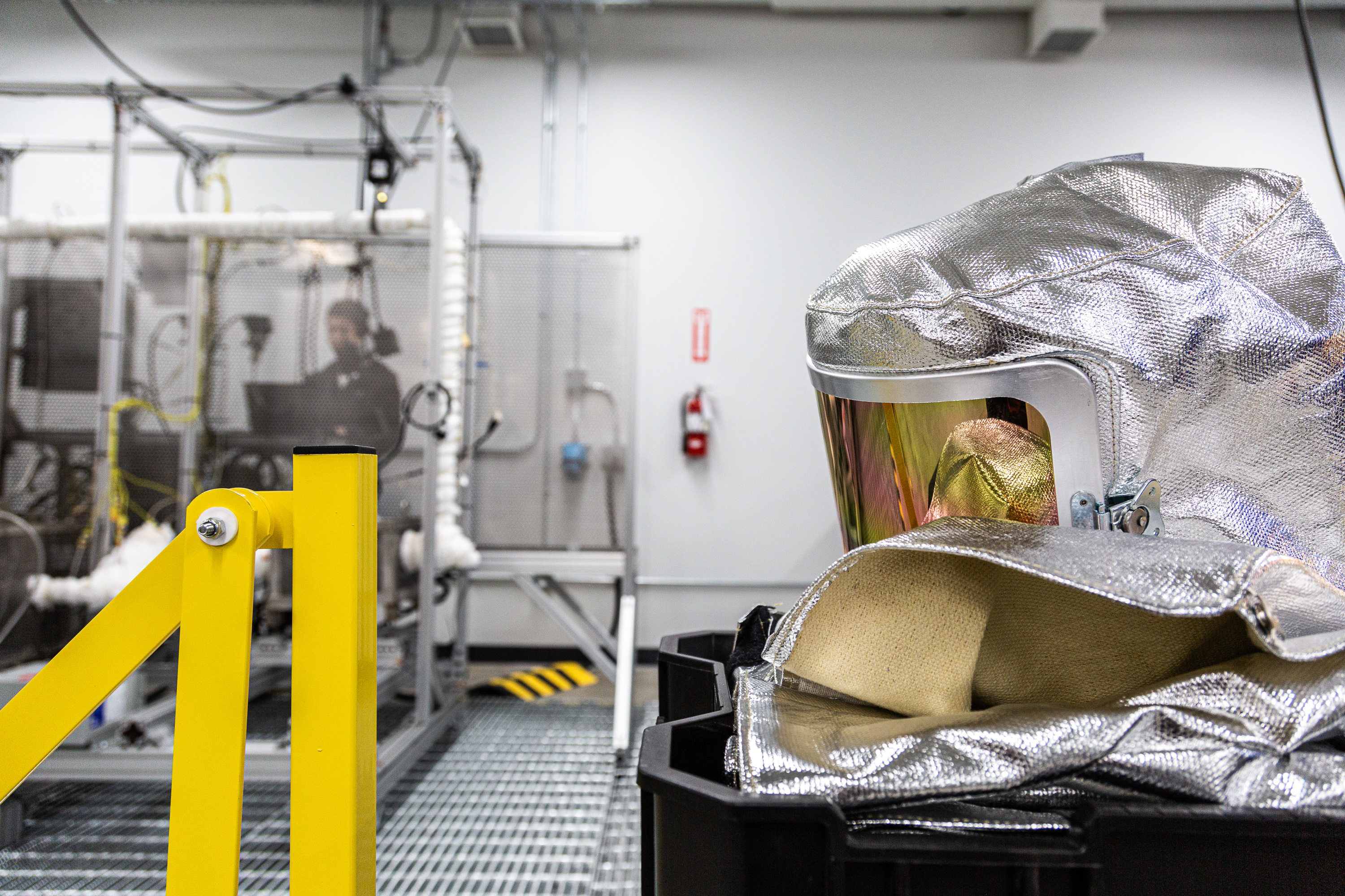 A silver safety hood with reflective visor and body suit are laid before a man within an industrial test stand.