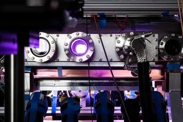 A purple light of a plasma radiates from the second of four circular flanges of a fusion device.