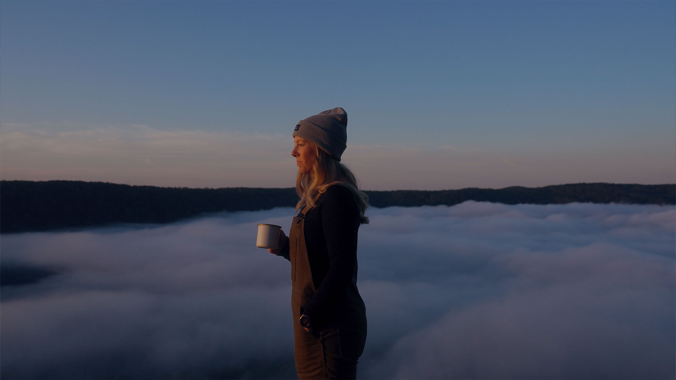 Sara holding a coffee mug on a mountaintop above the clouds at sunrise