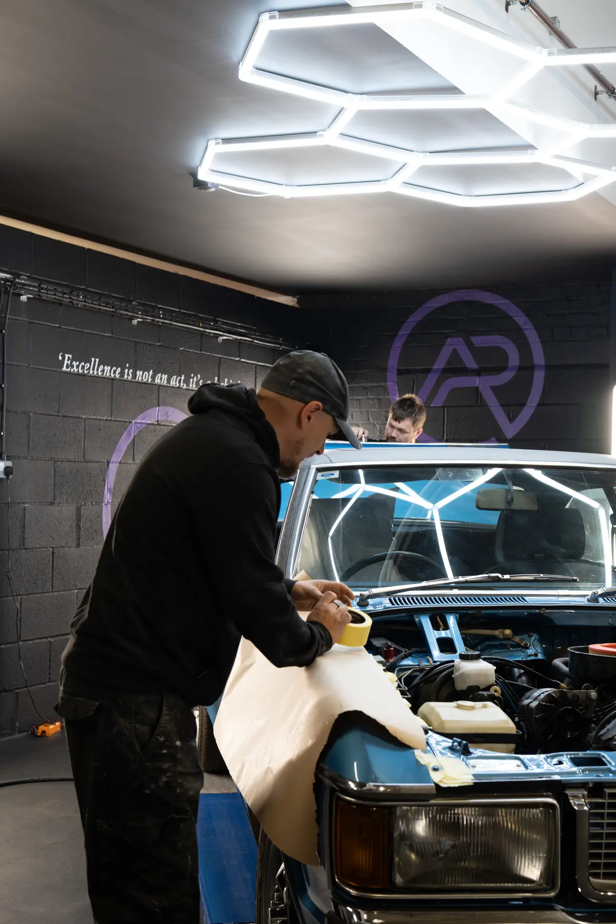 A team member applies masking paper to the front wing of the blue Ford Granada Coupé.