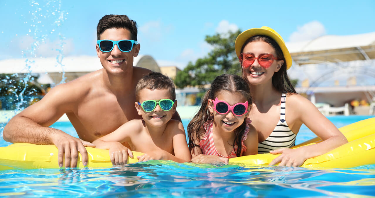 Padres y dos niños pequeños sonriendo con gafas de sol de colores sobre un inflable amarillo en una piscina azul brillante