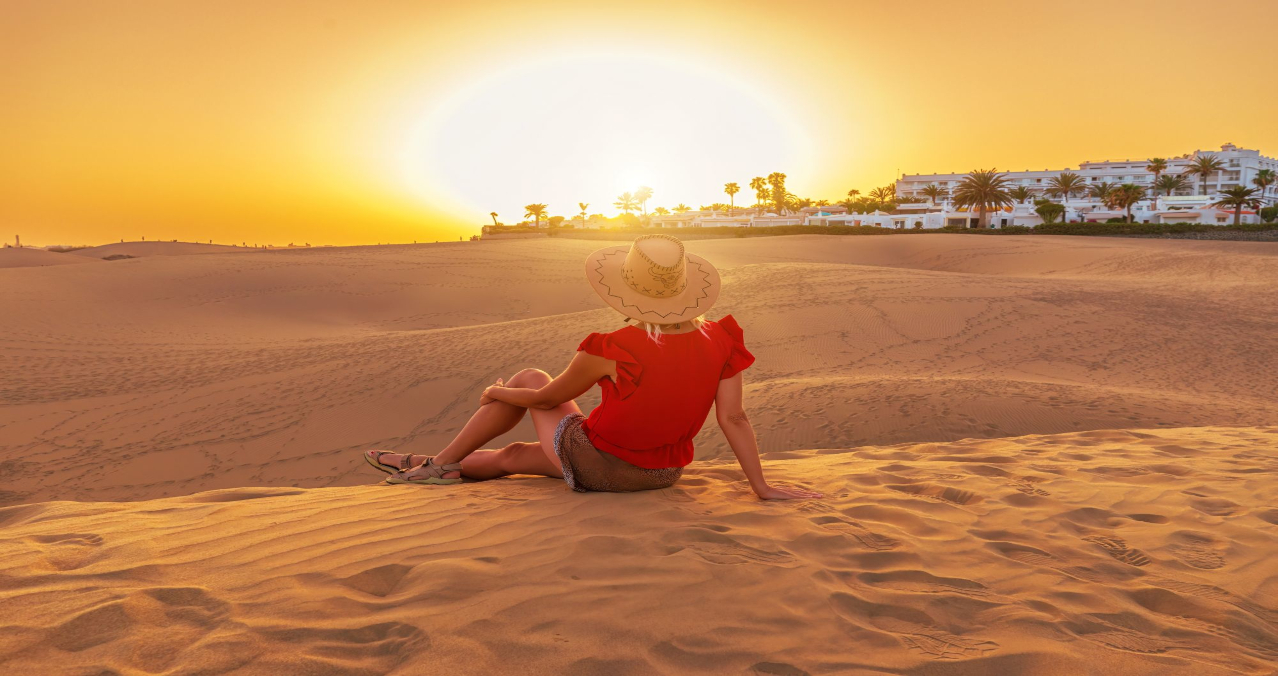 Viajera con sombrero sentada de espaldas contemplando un espectacular y cálido atardecer dorado sobre las dunas de Maspalomas