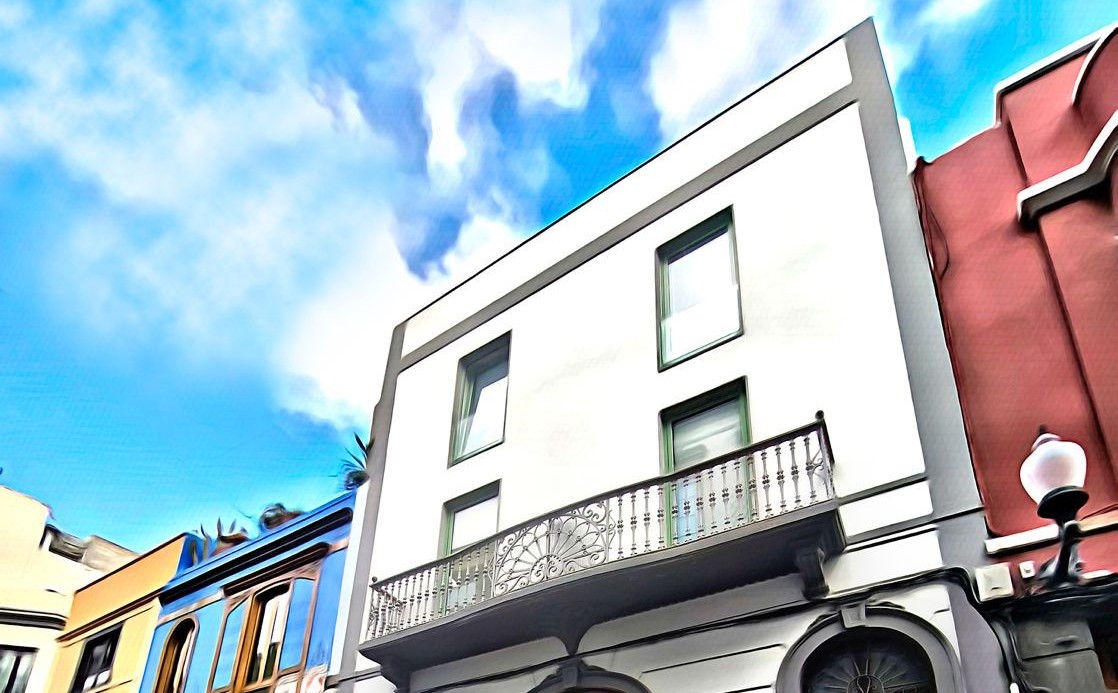 Plano contrapicado de un edificio blanco y elegante con balcón de hierro forjado rodeado de fachadas de colores tradicionales en el barrio de Triana
