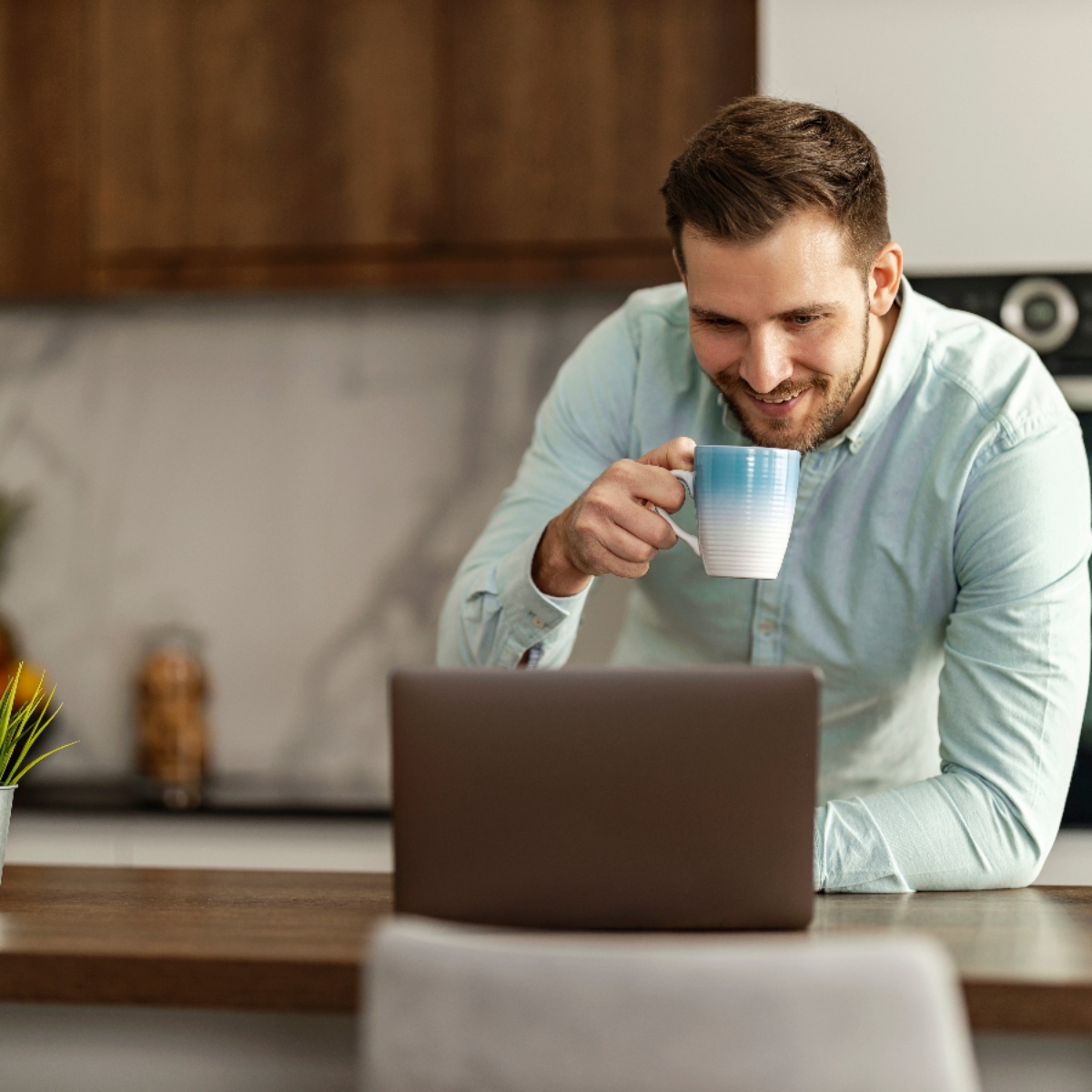 Nómada digital sonriendo mientras toma café y trabaja con su ordenador portátil en la cocina moderna de un apartamento vacacional