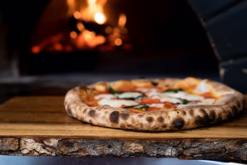 Primer plano de una auténtica pizza napolitana sobre tabla de madera rústica con un horno de leña encendido al fondo