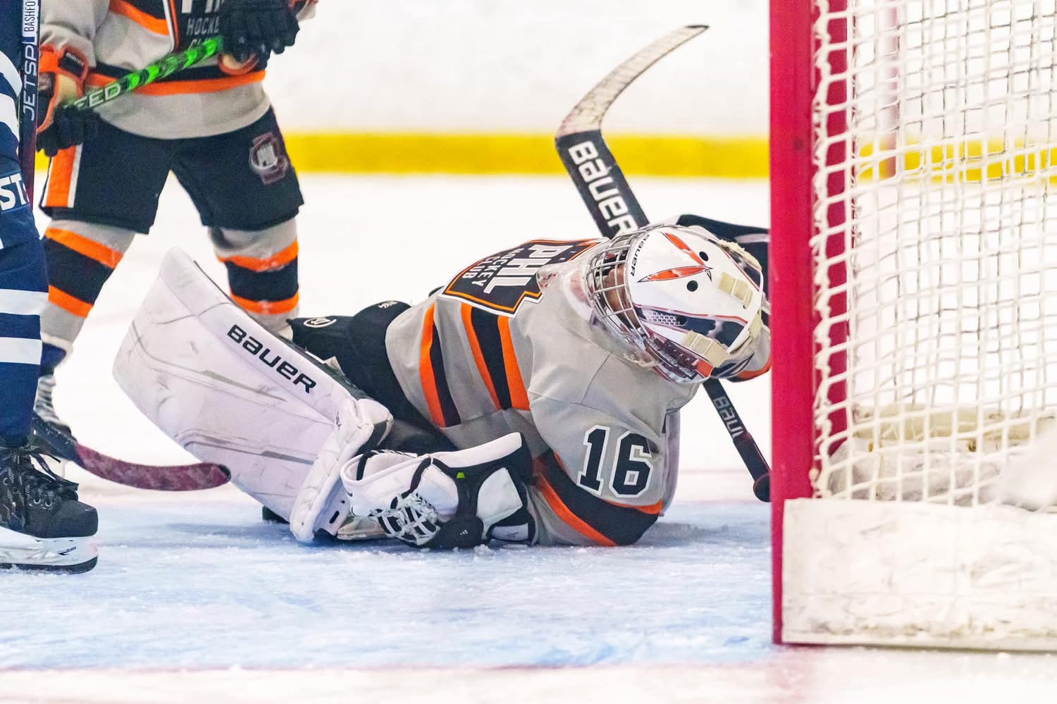 Youth goaltender in grey and orange equipment lies on the ice next to the right goalpost in game action