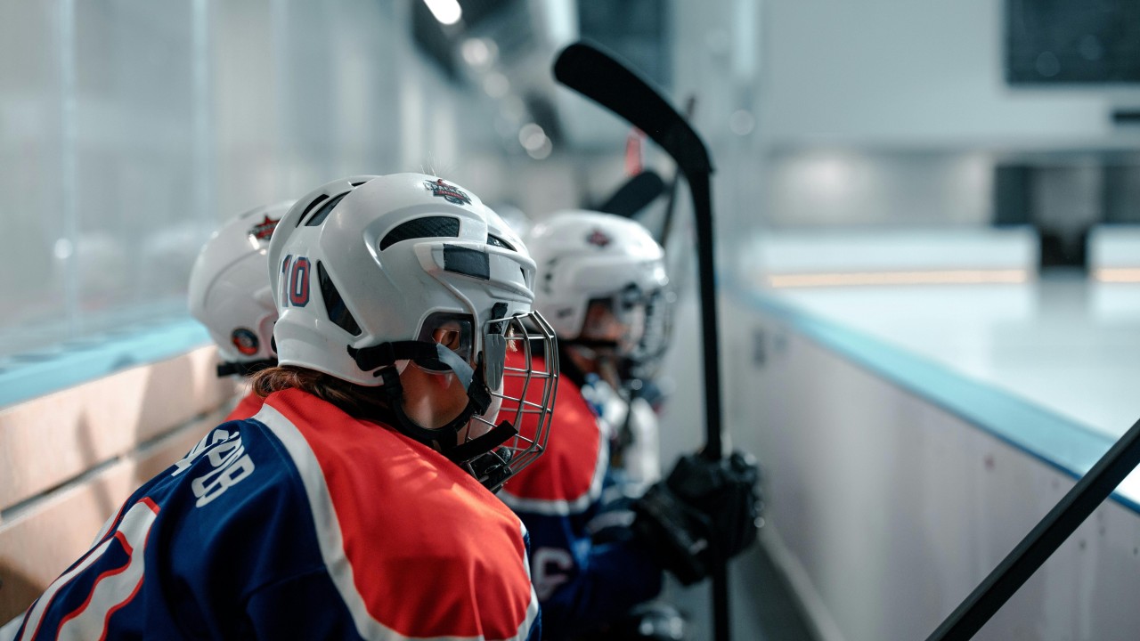 Youth hockey players in red, white, and blue uniforms watching the ice from the team bench.