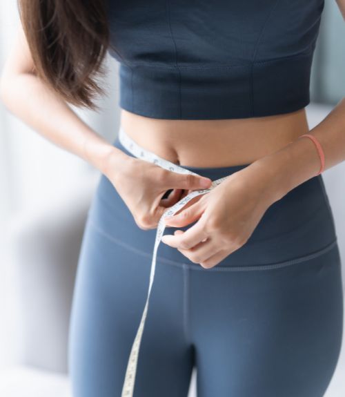 Woman in dark workout clothes measuring her waist with a tape measure.