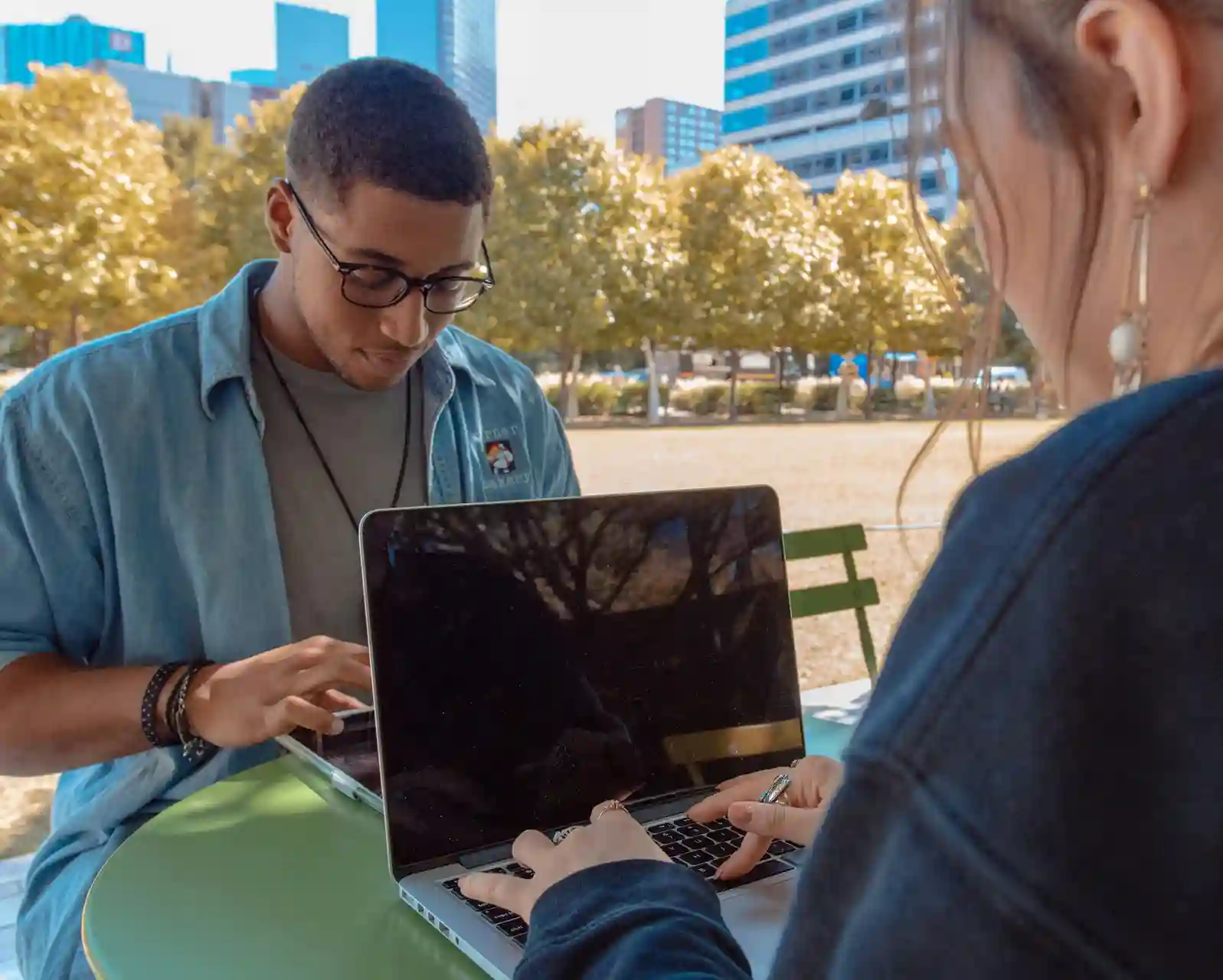 A young Black man wearing glasses and a denim shirt, and a woman viewed from over her shoulder, working on laptops together at a small green table outdoors. They are in a sunny urban park; autumn trees and modern skyscrapers are visible in the background.