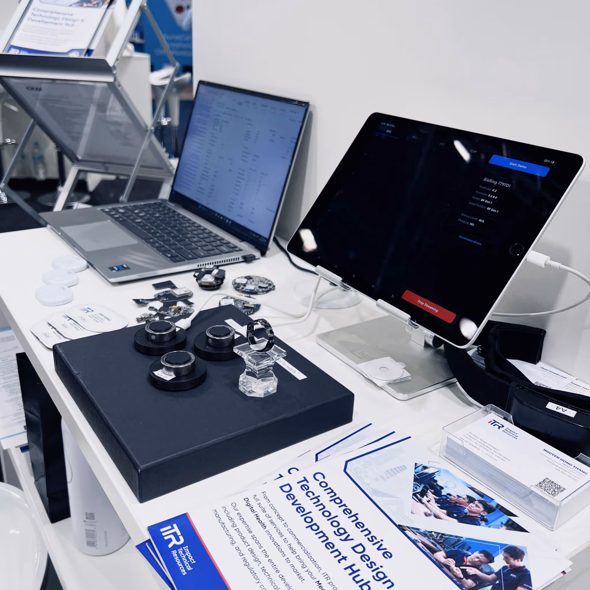 ITR workspace display featuring a silver laptop with a data spreadsheet, an iPad showing data editing options, and a central board with three innovative hardware prototypes surrounded by electronic components, brochures, and business cards. The setting highlights ITR’s expertise in comprehensive technology design and development for digital health, MedTech, and smart automation solutions.