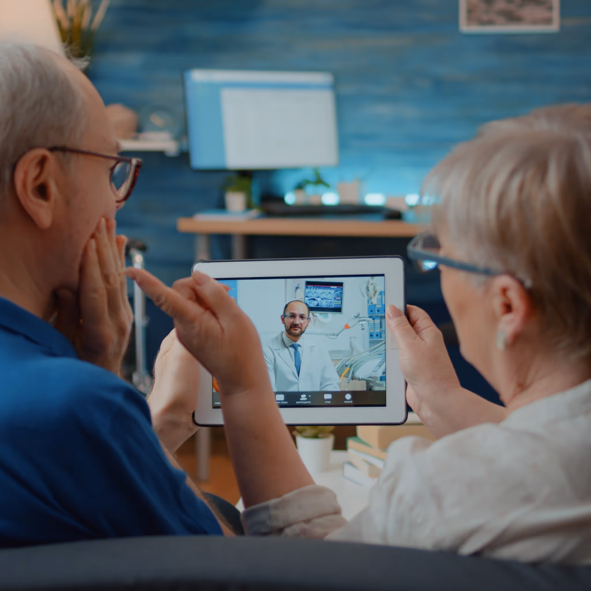 Elderly couple seated on a couch at home, engaged in a virtual consultation with a smiling doctor displayed on a tablet. The woman gestures while holding the device, and the man listens attentively. The scene highlights ITR’s digital health solutions, enabling accessible and comfortable telemedicine experiences in a modern, inviting home environment.