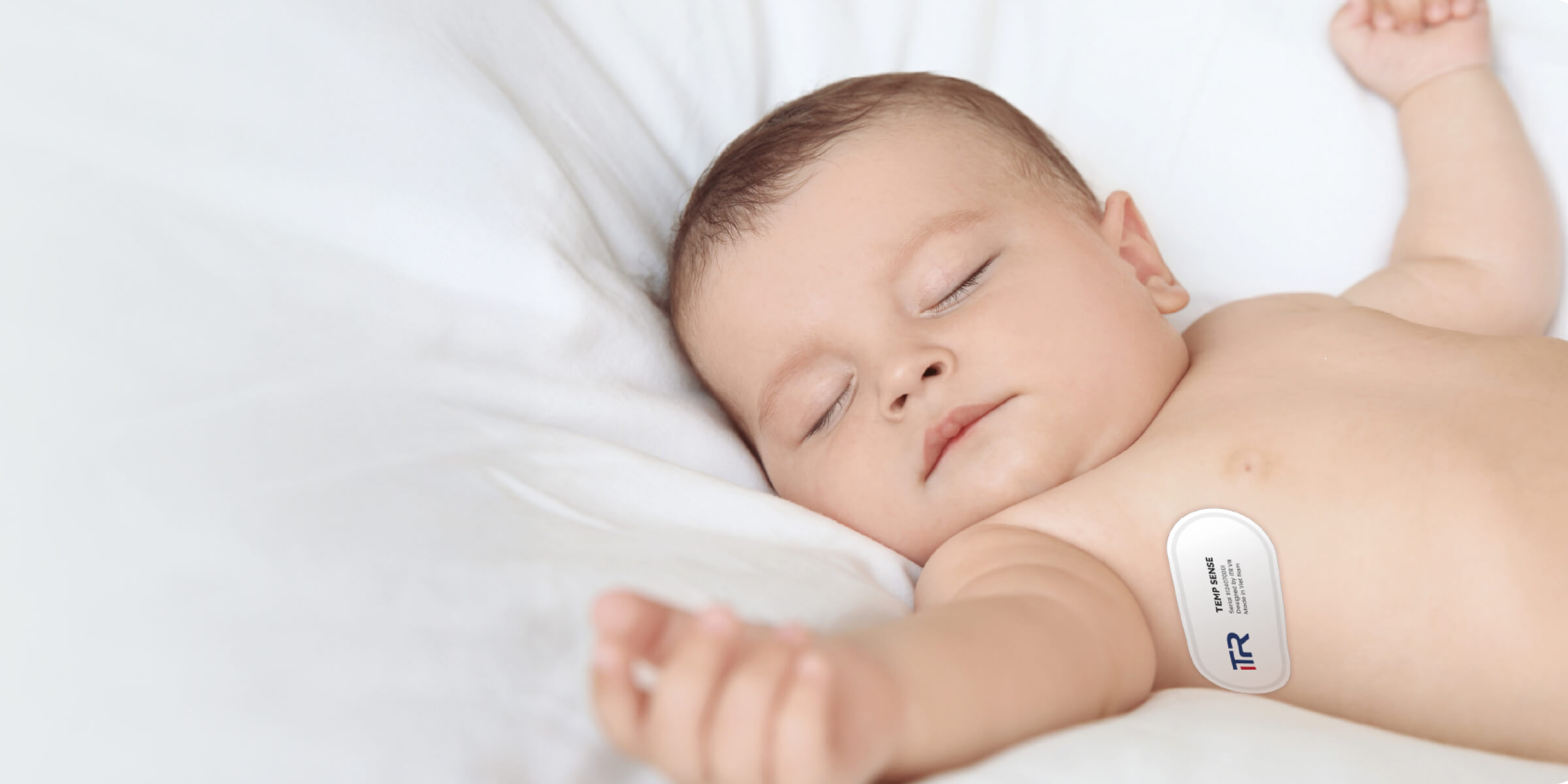 Sleeping baby lying on white bedding with a small medical sensor attached to the chest.