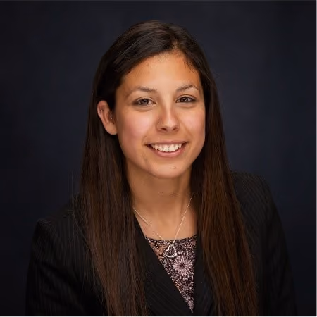 Smiling woman with long dark hair wearing a black blazer and a silver heart necklace on a dark background.