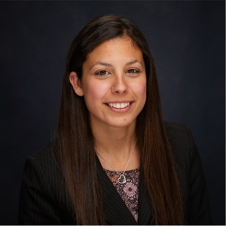 Smiling woman with long dark hair wearing a black blazer and a silver heart necklace on a dark background.