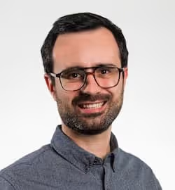 Smiling man with dark hair, beard, glasses, and button-up shirt on a white background.