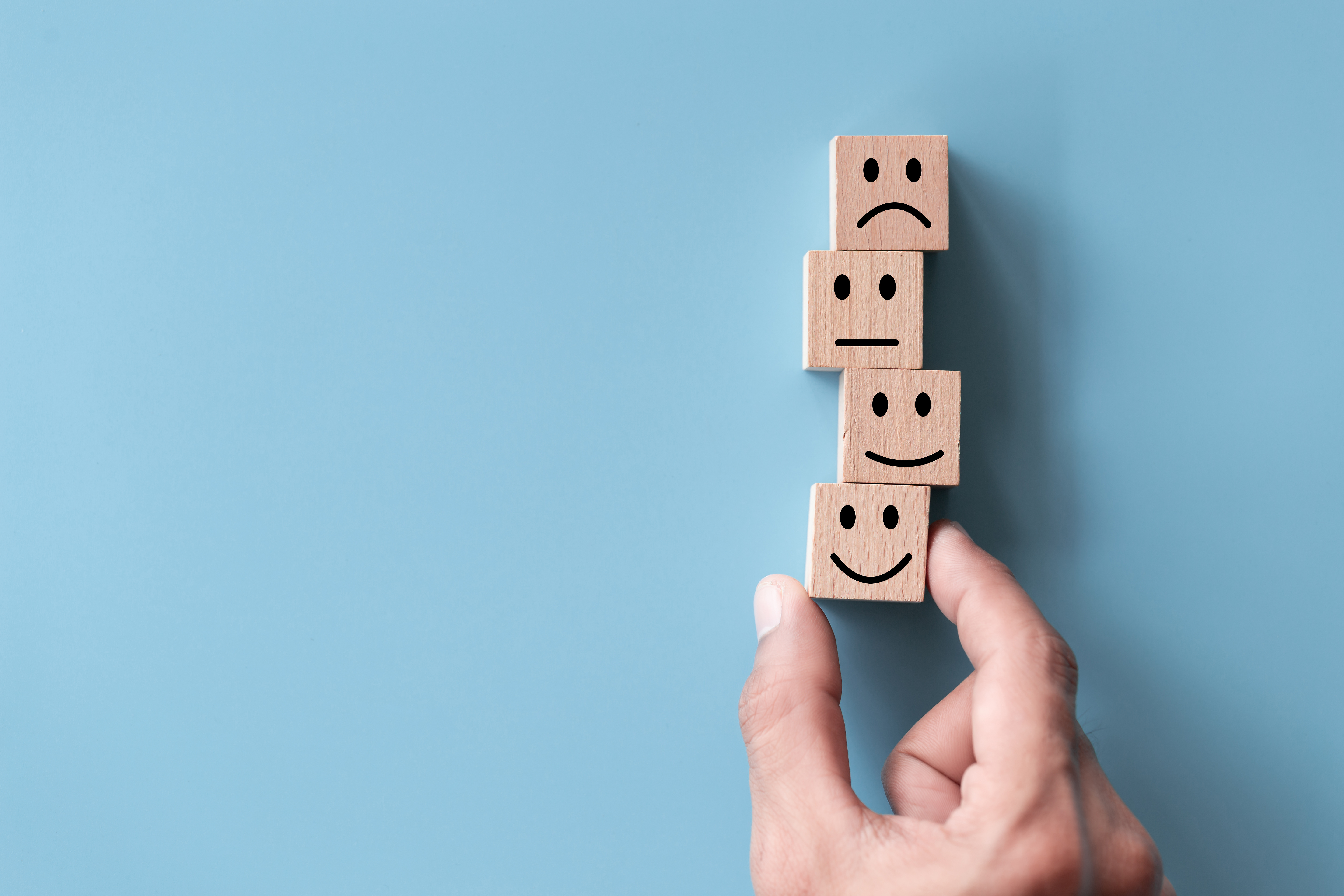 A photo with four wooden blocks stacked on top of each other on a blue background. Each wooden block has a smiley face on it, increasing in satisfaction.